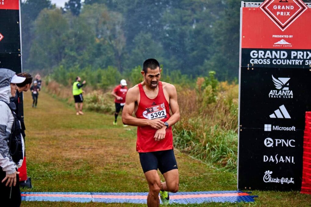 A man just crossed the finished line of a cross country race, stopping his watch, and his face is in pain.